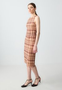 Frau in einem ärmellosen, mittellangen beigen Kleid mit braunem Karomuster und schwarzen Absatzsandalen vor einem schlichten, hellen Hintergrund.