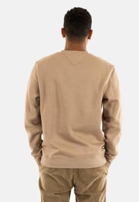 Sudadera beige con cuello redondo, tejido texturizado y detalle de costura en V en la parte posterior, combinada con pantalones de pana.