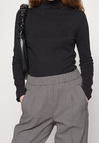Woman wearing black turtleneck sweater, grey houndstooth high-waisted trousers, with hands in pockets and black ruched leather shoulder bag.