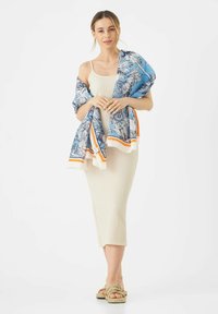 Robe midi en tricot beige avec une finition texturée, superposée avec un châle floral bleu aux accents orange, et accompagnée de sandales à brides en corde.