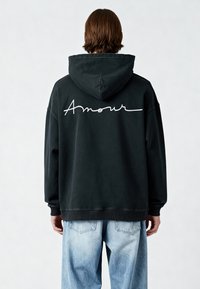 Felpa nera con cappuccio caratterizzata da un ricamo bianco con la scritta "Amour" sul retro. Il tessuto è morbido al tatto, con polsini e orlo a coste.