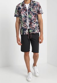 Muž v košili s krátkým rukávem s květinovým vzorem, bílem tričku, černých šortkách a bílých teniskách stojí na bílé podlaze.