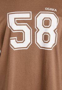 Rjava bombažna majica s potiskom belih blok številk "58" in napisom "OSAKA" nad njimi. Tkanina je gladka z rahlim sijajem.