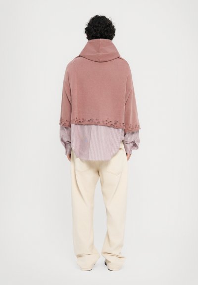 Oversized pink cotton hoodie with a front hem featuring cutout detailing, layered over a striped shirt and cream wide-leg pants.