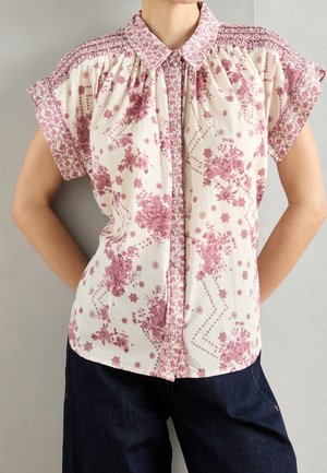 Nainen, jolla on lyhythihainen, napitettava pusero, jossa on vaaleanpunaisia kukkakuvioita ja rypytetyt olkapäät, yhdistettynä tummiin farkkuihin.