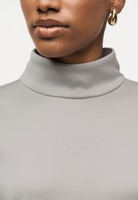 Femme portant un col roulé gris clair et une boucle d'oreille dorée en forme de goutte, avec une peau lisse et une couleur de lèvres neutre.