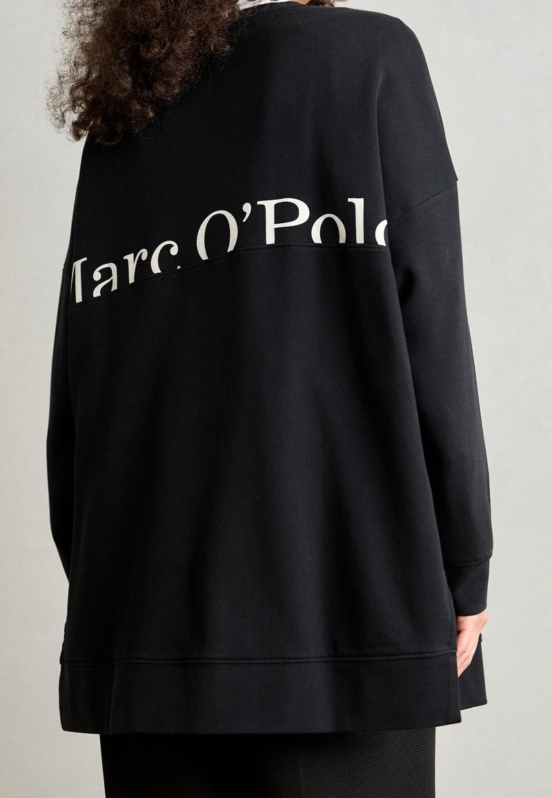Personne portant un sweat-shirt noir ample avec du texte blanc "Marc O'Polo" sur la partie haute du dos et des cheveux bouclés visibles.