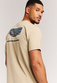 Beige T-shirt met korte mouwen en een grafische print van bergen en een zon in blauw, groen en wit op de achterkant. Soepele textuur.