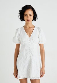 Biele krátke šaty s puff rukávmi, ktoré majú očkový vzor, výstrih do V a predné zapínanie na gombíky. Hladká látka s priliehavým pásom a rozšíreným lemom.