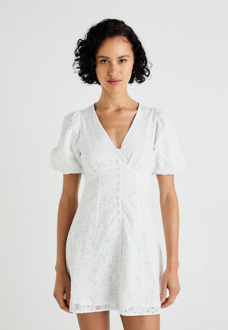 Biele krátke šaty s puff rukávmi, ktoré majú očkový vzor, výstrih do V a predné zapínanie na gombíky. Hladká látka s priliehavým pásom a rozšíreným lemom.