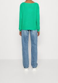 Grön stickad tröja med bred halsringning, långa ärmar och ribbade mudd, kombinerad med ljusblå högmidjade jeans och klumpiga vita skor.