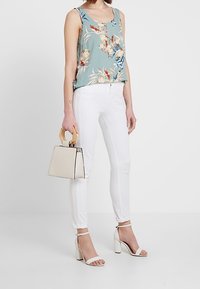 Top de tirantes floral en azul claro con patrones en blanco y verde, combinado con jeans blancos ajustados y sandalias de tacón beige. Bolso pequeño color crema con asas de madera.