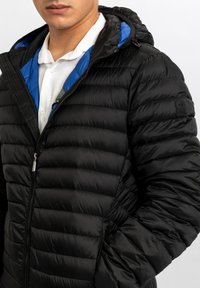 Giacca a vento nera imbottita con fodera blu, chiusura anteriore con cerniera e cappuccio. Presenta una trama trapuntata e un logo ricamato sulla manica.