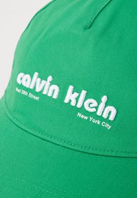 Cappello in cotone verde con ricamo bianco che dice: "calvin klein" e "West 39th Street, New York City." Il testo è in grassetto e di grande impatto.