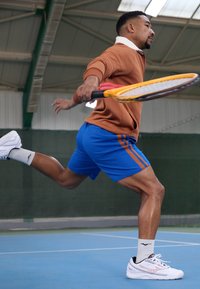 Atelet pruunis pikkade varrukatega särgis ja sinistes lühikestes pükstes, serveerib tennisepalli kollase reketiga, jalas valged ja mustad spordijalatsid.