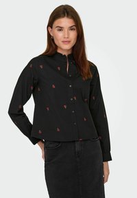 Donna che indossa una blusa nera con bottoni e motivi floreali rossi, insieme a una gonna di jeans nera, in piedi davanti a uno sfondo chiaro e semplice.