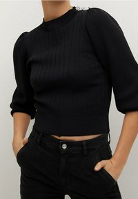 Haut en maille côtelée noir avec un col montant, des manches bouffantes et un accent bijou sur l'épaule. Associé à un pantalon noir taille haute.