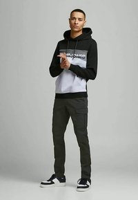 Junger Mann steht mit gefalteten Händen, trägt einen schwarzen und grauen Jack & Jones Kapuzenpullover, dunkle Cargo-Hosen und schwarz-weiße Sneakers vor grauem Hintergrund.