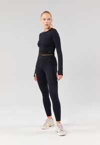 Schwarzes Crop-Top mit langen Ärmeln und hoch taillierte Leggings aus glattem Stretchstoff. Füße in hellen Sportschuhen.