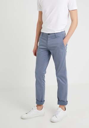 Persoon draagt lichtblauwe opgerolde broek, witte sneakers en een wit T-shirt met korte mouwen, staat met één hand in de zak.