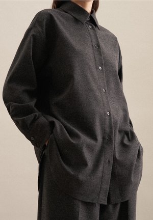 Osoba ubrana w luźną, ciemnoszarą koszulę na guziki z kołnierzykiem i długimi rękawami, ręce w kieszeniach, na jednolitym beżowym tle.