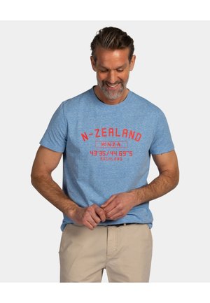 Man glimlacht naar beneden, draagt een blauw "N-ZEALAND AUCKLAND" T-shirt en beige broek, vingers aanpassend tegen een effen lichte achtergrond.