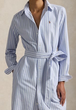 Femme portant une robe-chemise à rayures verticales bleu clair et blanches, manches longues, col, boutons sur le devant et ceinture nouée à la taille.