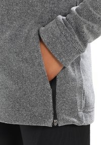 Ręka w bocznej kieszeni szarej polarowej kurtki z widocznym czarnym suwakiem i detalami mankietów na białym tle.