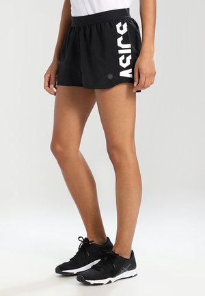 Frau trägt schwarze Asics Laufshorts und schwarze Sportschuhe, steht vor einem schlichten hellen Hintergrund.