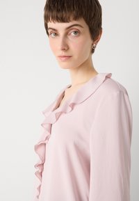 Vaaleanpunainen paita, jossa on röyhelökaulus ja pitkät hihat, valmistettu pehmeästä kankaasta, joka on sileä ja miellyttävä. Korostuu hienovaraisella kiillolla.