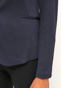 Manche et détail de poche d'un blazer texturé bleu marine porté avec un pantalon noir et une main visible sur le côté.