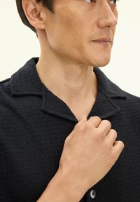 Czarna teksturowana koszula z kołnierzykiem, zapinana na guziki, z subtelnym wzorem. Zbliżenie na rękę regulującą kołnierzyk.