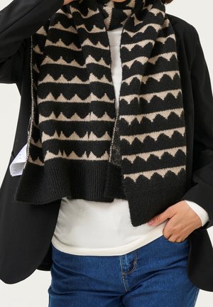 Personne portant une écharpe à motifs géométriques noirs et beiges, chemise blanche, veste noire et jean bleu, avec une main dans la poche.