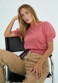 Maglione corto rosa con maniche a sbuffo e pantaloni beige a vita alta dotati di una cintura. Modella seduta su una sedia nera.