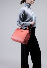 Koraal lederen handtas met gestructureerde vorm, dubbele bovenhandvaten en discreet merklogo. Draagt door een persoon in een grijze coltrui en zwarte broek.