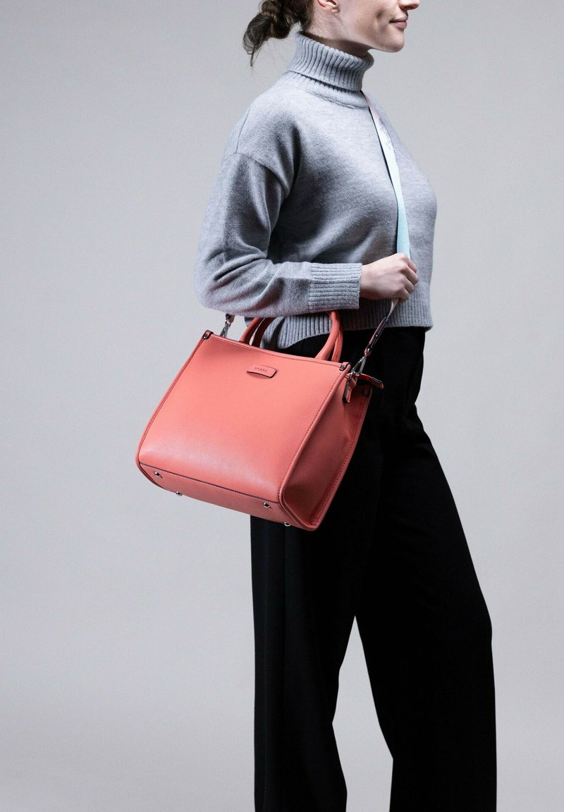 Koraal lederen handtas met gestructureerde vorm, dubbele bovenhandvaten en discreet merklogo. Draagt door een persoon in een grijze coltrui en zwarte broek.