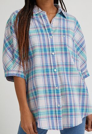Woman wearing loose button-up shirt with pastel blue, green, and purple plaid pattern and chest pocket.