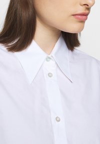Vit skjorta med stor krage, försedd med en rad knappar framtill. Slät textur och klassisk design som framhäver subtila knappdetaljer.