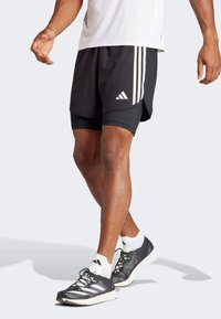 Pantalones cortos deportivos negros con tres rayas blancas y logo, que cuentan con un ajuste relajado y un diseño ligero, combinados con zapatillas de correr negras.