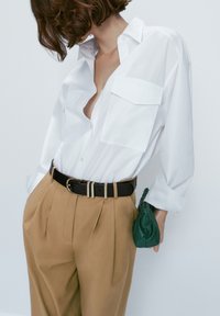 Vit skjorta med knappar och två bröstfickor, kombinerad med högmidjade, tanfärgade, veckade byxor och en liten grön väska. Svart bälte med silverspänne.