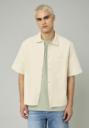 Chemise à manches courtes, col, en tissu crème texturé, avec une poche devant. Portée par-dessus une chemise vert clair, associée à un jean bleu.