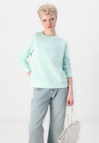 Gai minta zaļš sporta krekls ar reljefu "GANT" logo, ribotām piedurknēm un apakšmalu, kombinēts ar gaiši ziliem platiem džinsiem un bāli pelēku rokassomu.