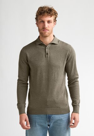 Mann mit lockigem Haar, der ein olivgrünes langärmeliges Poloshirt und blaue Jeans trägt, steht vor einem schlichten hellen Hintergrund.