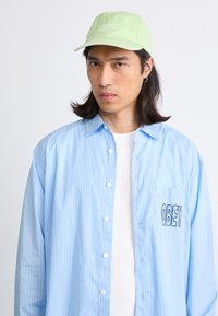 Camisa de rayas azul claro con un bolsillo que lleva bordado "OBEY", superpuesta sobre una camisa blanca. Gorra verde claro con texto.