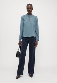 Lichtblauwe blouse met een gerimpelde halslijn en sleufdetail, gecombineerd met marineblauwe wijde broekspijpen en puntige bruine schoenen. Accessoires: zwarte handtas.
