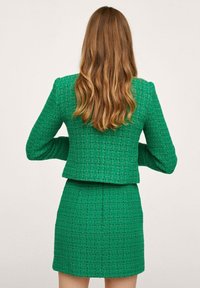 Vihreä tweed-setti, johon kuuluu lyhyt takki jämäkkäolkkien kanssa ja tiukka mini-hame, joissa molemmissa on teksturoitu neliökuvio.
