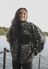 Faux fur-jacka med leopardmönster i svart, tan och kräm, med en svart ficka med dragkedja och hög krage.