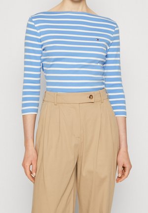Femme portant un haut à manches 3/4 rayé bleu et blanc, rentré dans un pantalon plissé beige taille haute avec une taille boutonnée.