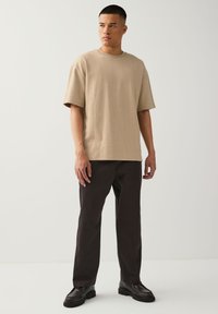 Man standing wearing beige short-sleeve textured shirt, black pants, and black chunky shoes, looking slightly to the left on a white background.