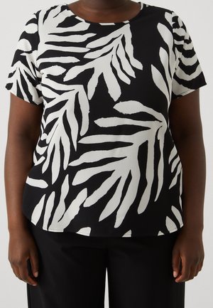 Sort og hvid kortærmet top med abstrakt bladmønster båret med sorte bukser på en person mod en ensfarvet baggrund.